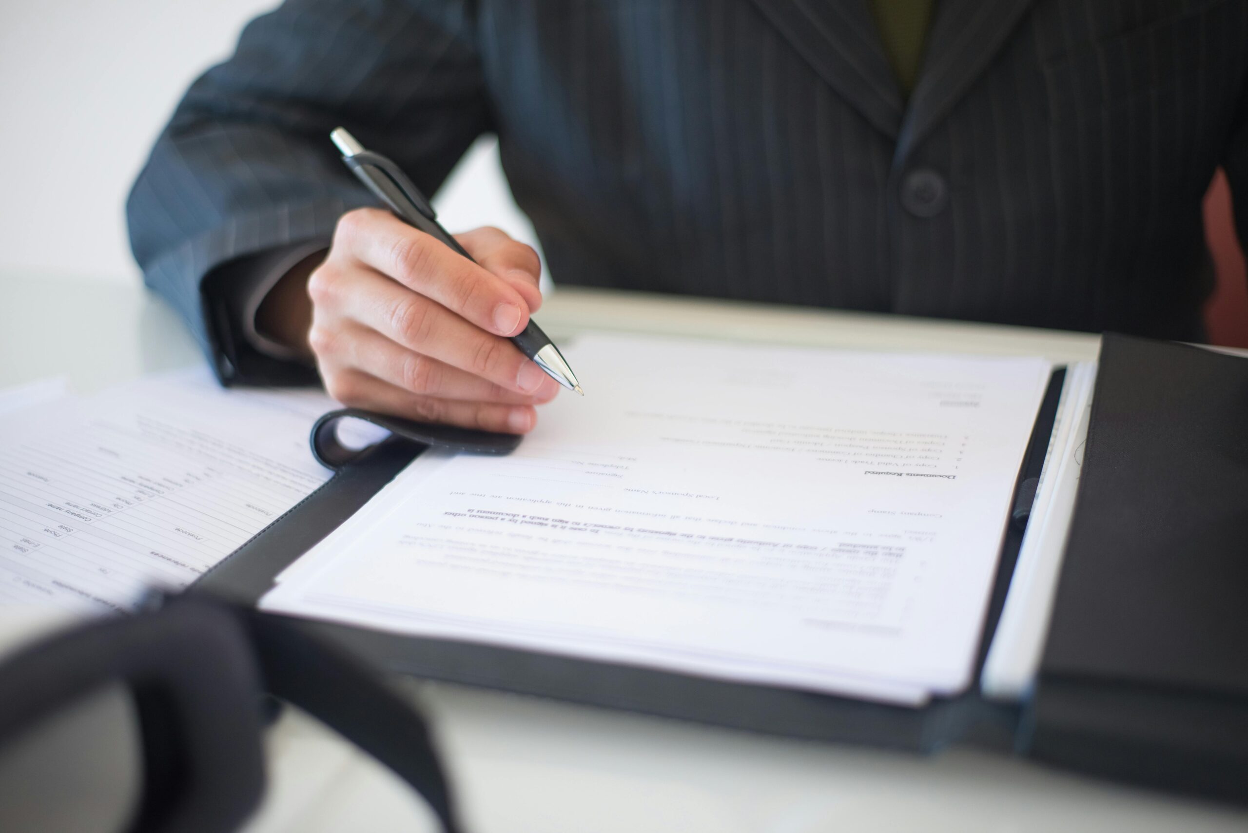 Person in a suit signing an application form held in a leather folder at a desk