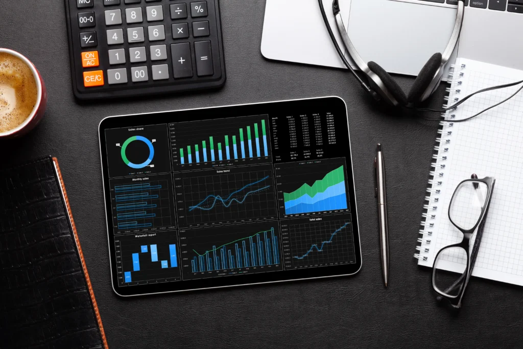 a top-down view of a tablet displaying financial charts and graphs on a dark office desk next to a calculator, notebook, and glasses