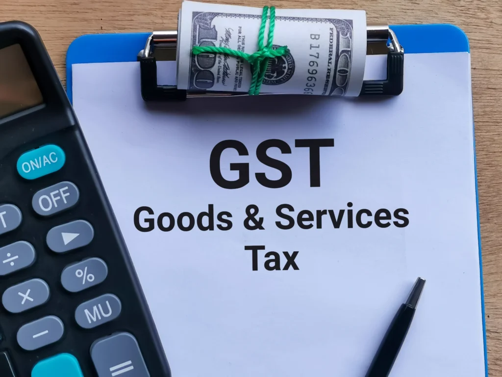 a clipboard holding a goods and services tax document and rolled currency next to an office calculator