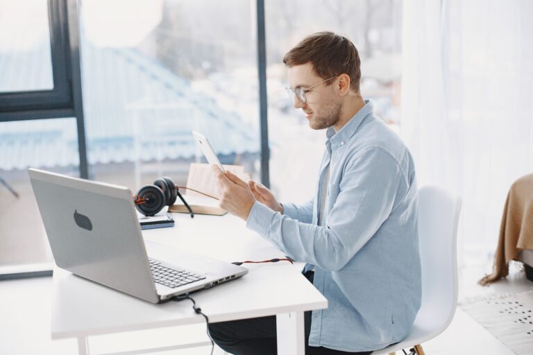 a man with glasses smiling while working with a tablet and laptop at a bright white desk