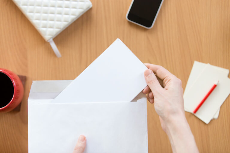 top-down view of hands opening a white envelope on a wooden office desk
