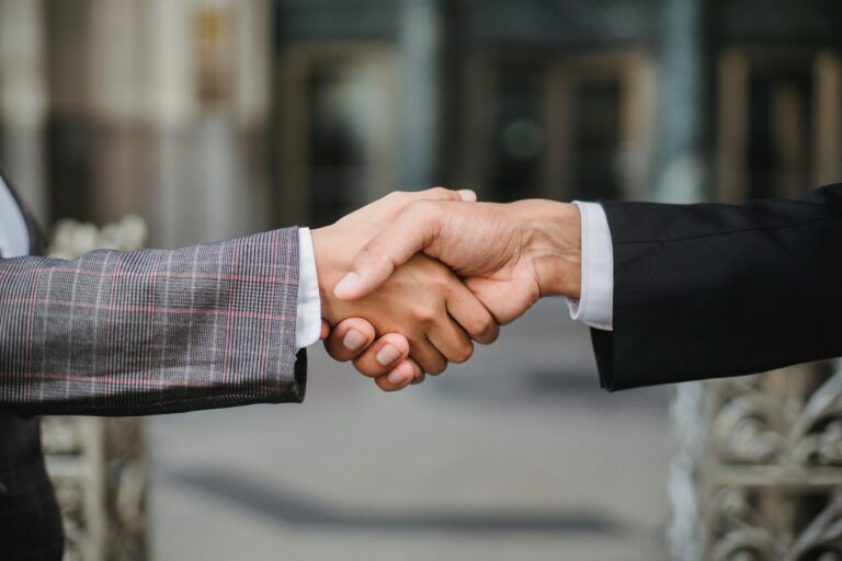 Two people in business suits shaking hands outside an office building, symbolizing a partnership or agreement